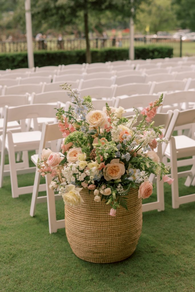 Virginia Beach wedding florist arrangement of peach garden roses, blush snapdragons, and blue delphinium in a woven basket beside white ceremony chairs