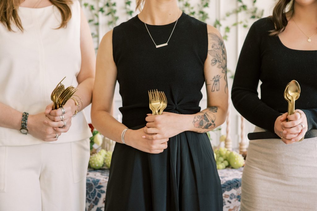 Three women in black and ivory outfits holding bundles of gold forks and spoons in front of a table set with a blue and white floral tablecloth, green grapes, and an ivy-covered wall
