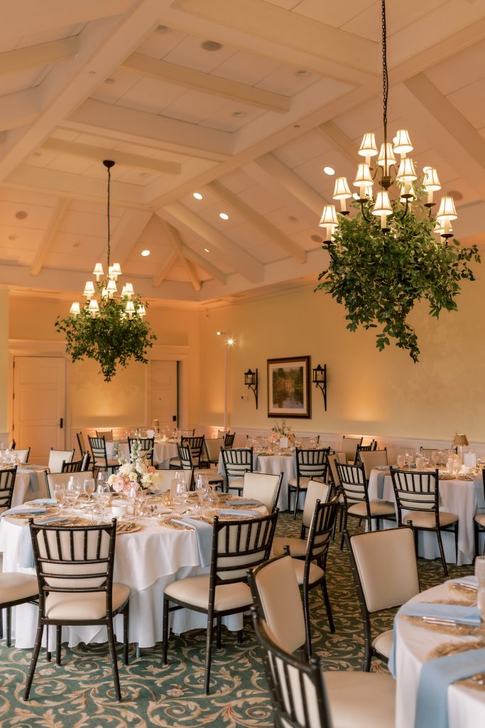 Wedding reception with round tables set in white tablecloths, dusty blue napkins, and woven chargers, black chiavari chairs, low blush and ivory centerpieces, and chandeliers wrapped in lush greenery by Virginia Floral Design & Events