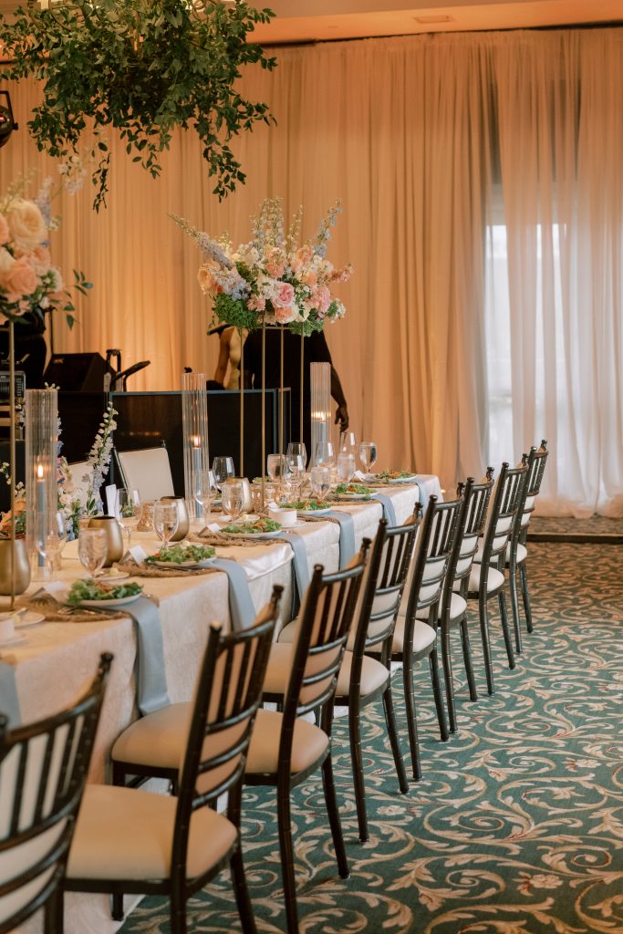 Long reception table with white tablecloths, dusty blue napkins, woven chargers, black chiavari chairs, and tall blush and blue floral arrangements on gold stands by Virginia Floral Design & Events