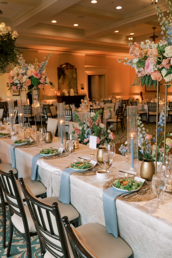 Wedding reception head table with dusty blue napkins, woven chargers, taper candles in glass hurricanes, and tall blush and blue floral arrangements by Virginia Floral Design & Events
